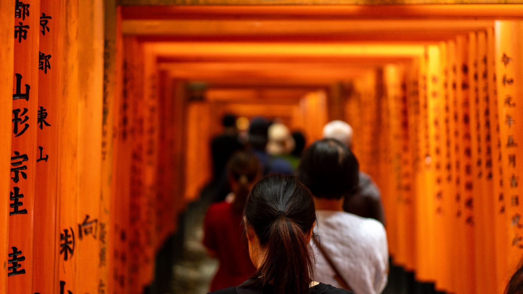 A woman with a ponytail walks through the vermilion Torii gates at Fushimi Inari, centered between the columns.